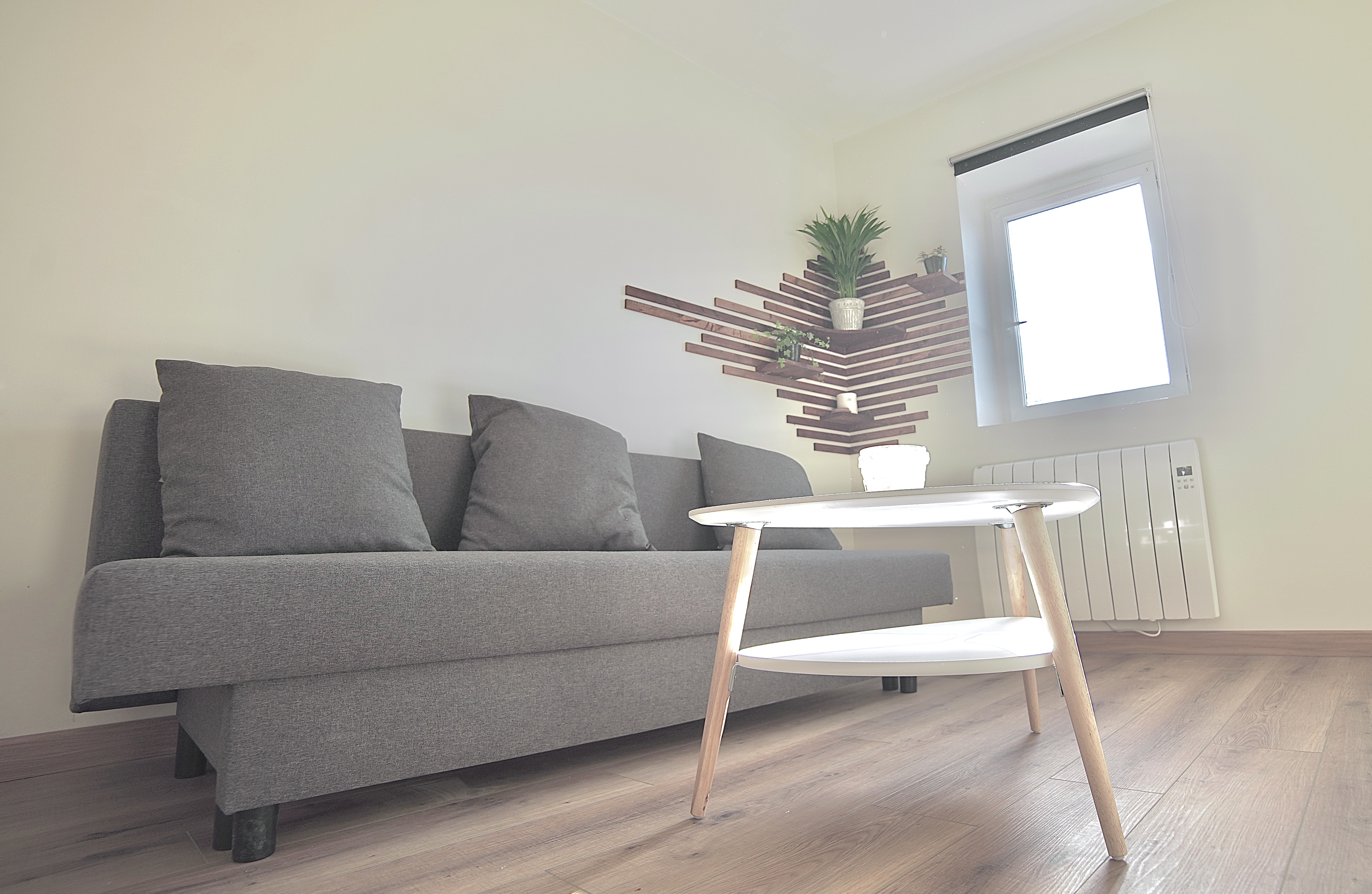 Salon avec canapé gris, table basse blanche sur parquet et étagère murale en bois avec plantes.