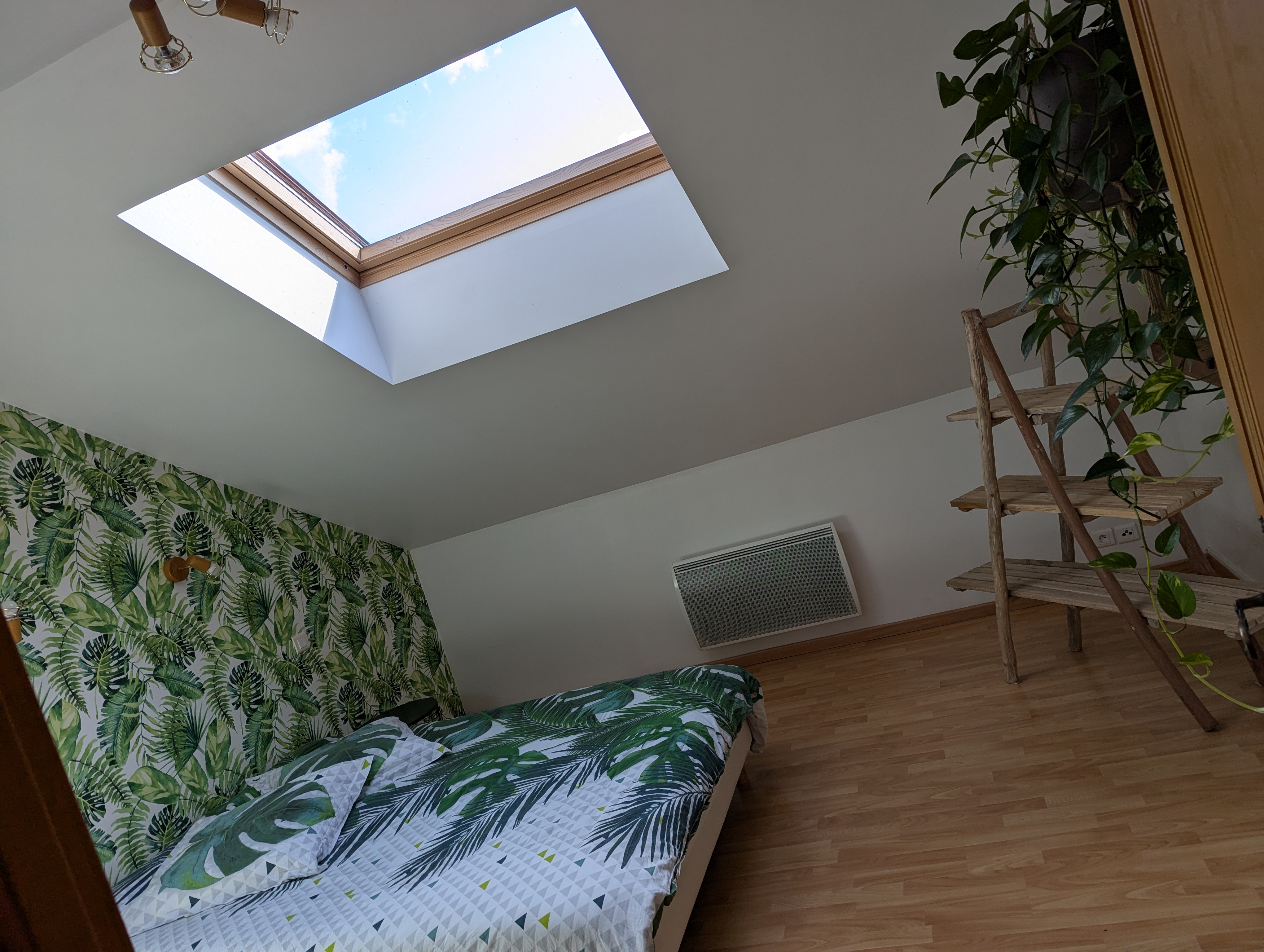 Chambre mansardée avec velux, lit double, papier peint feuilles vertes, radiateur électrique et étagère en bois.