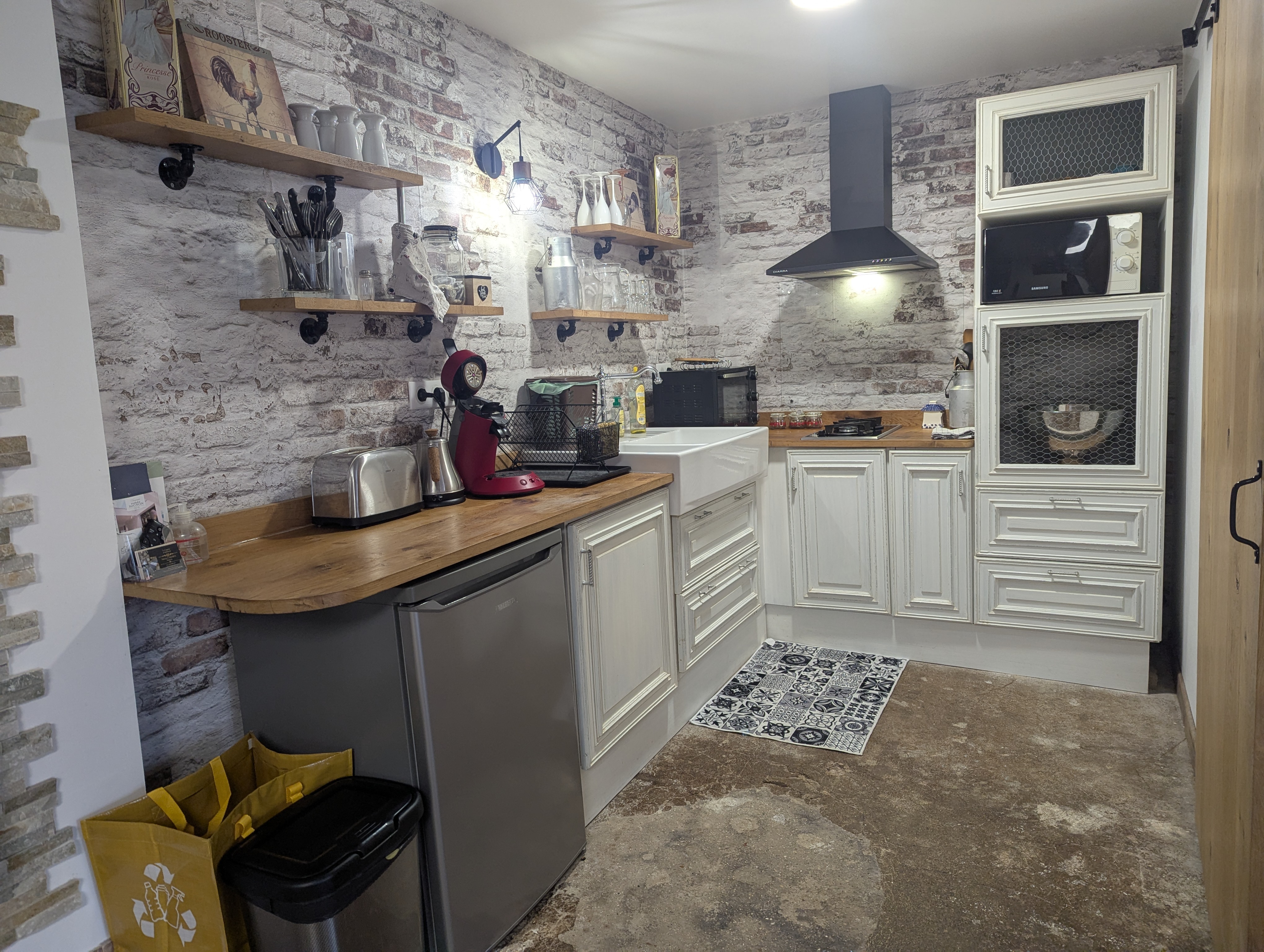 Cuisine équipée avec mur en brique apparente, plan de travail bois, hotte, évier à cuve et machine à café.