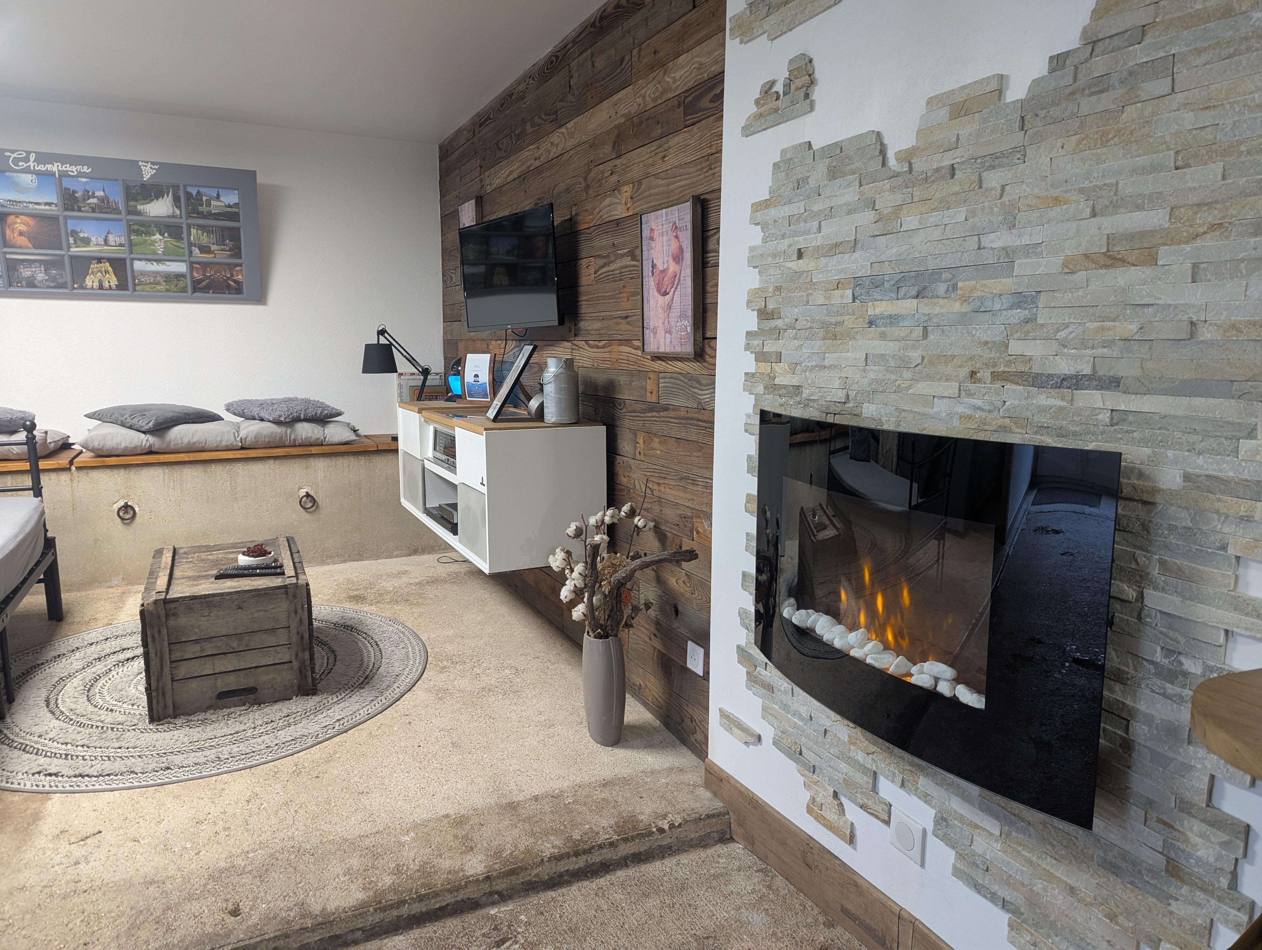 Coin salon avec cheminée électrique allumée, mur en bois de récupération, téléviseur et table basse en caisse en bois.