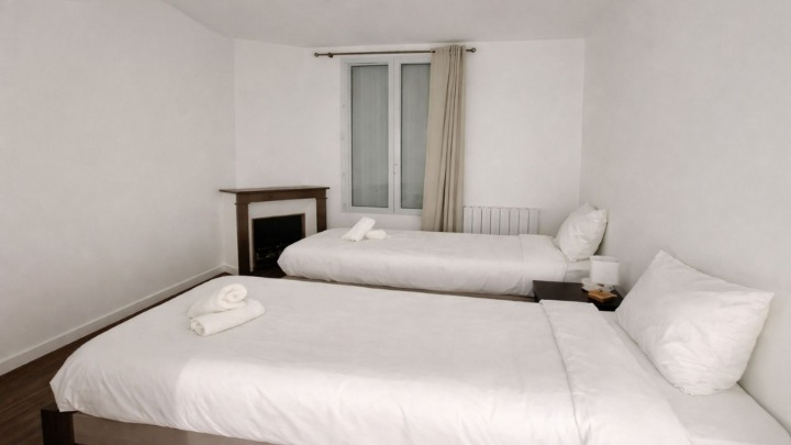 Chambre blanche avec deux lits simples en literie blanche, cheminée et radiateur.