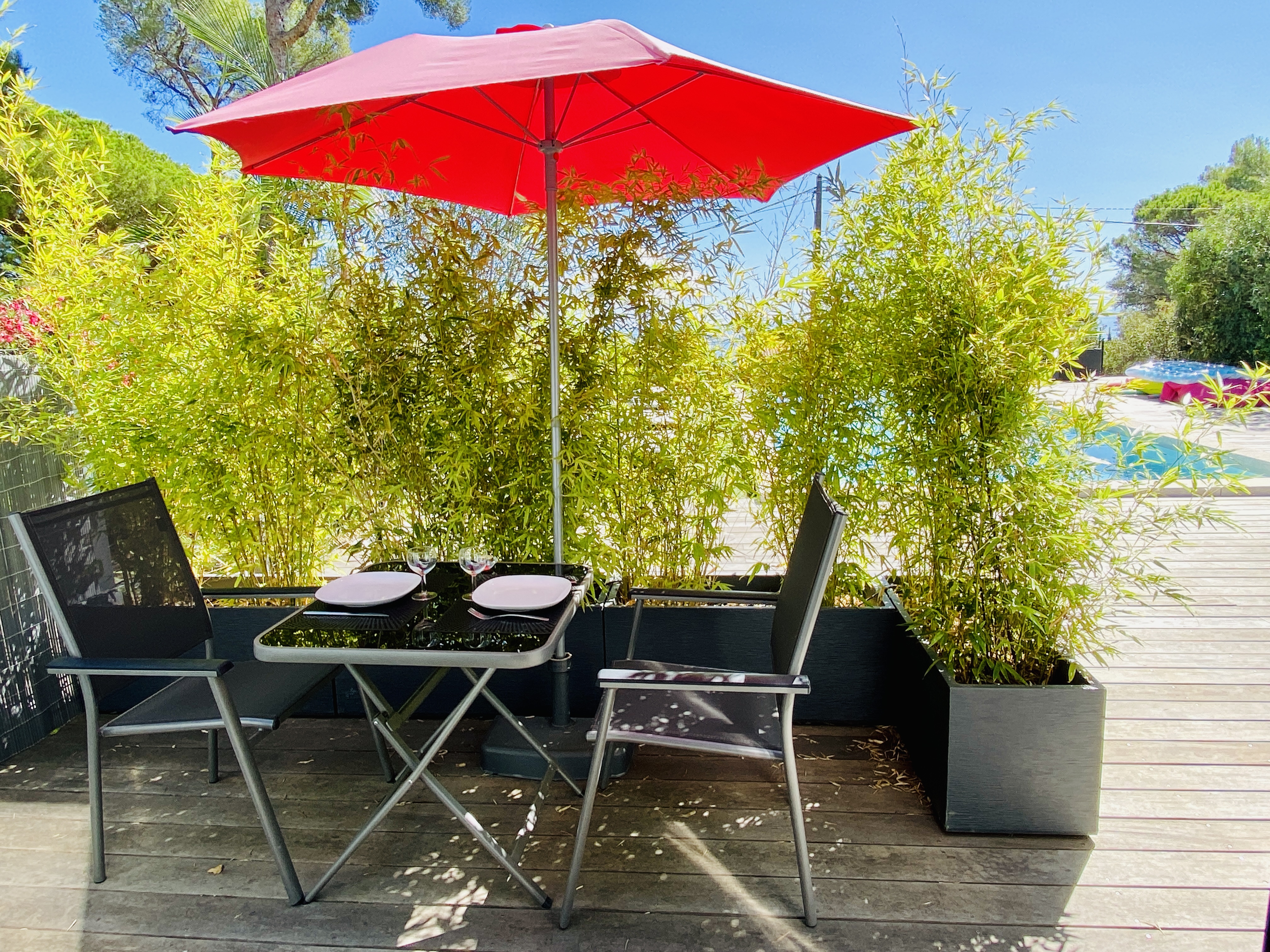 Terrasse en bois avec table, deux chaises et parasol rouge, vue sur piscine et jardins.