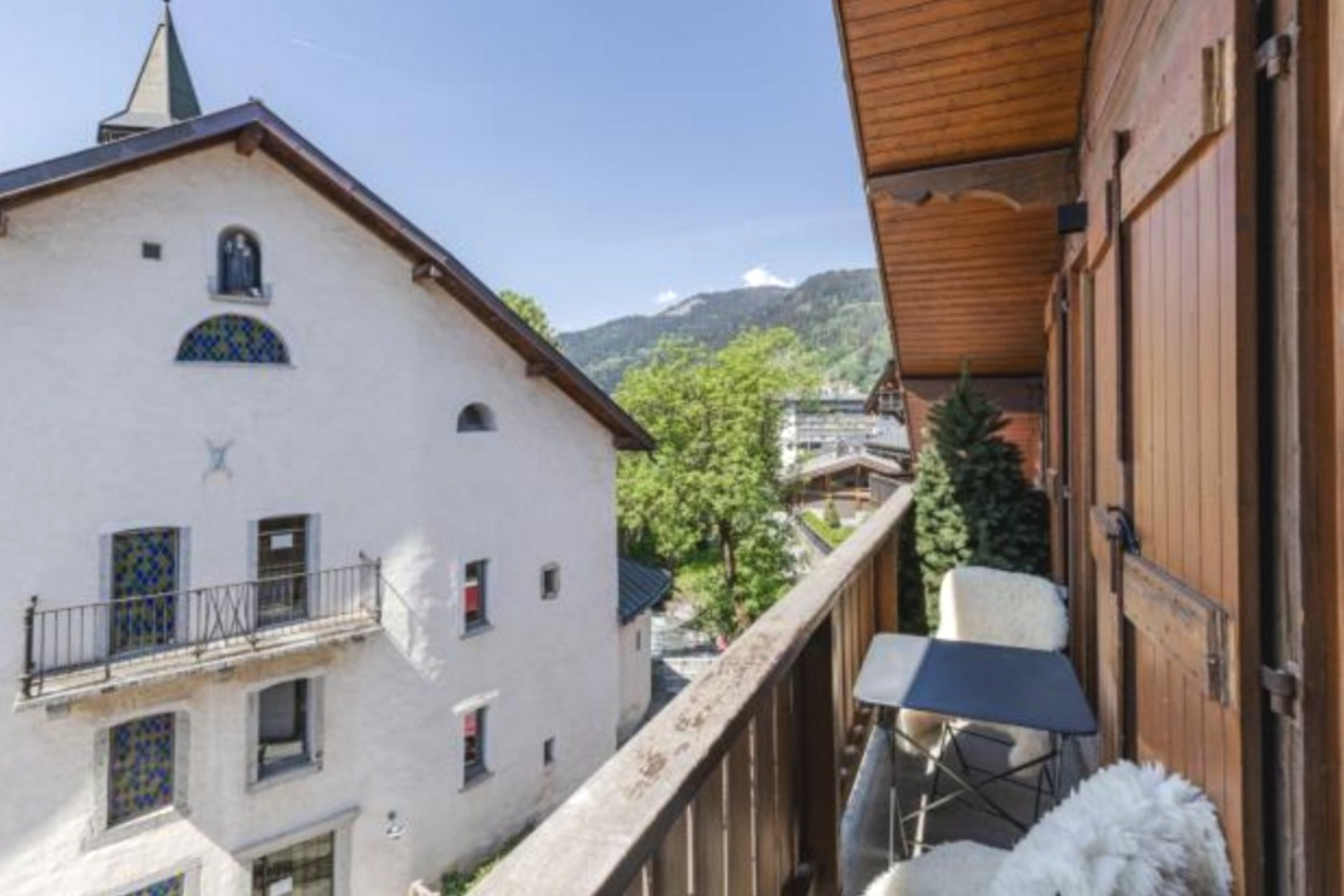 Balcon en bois avec chaise, vue sur bâtiment en pierre et massif montagneux.